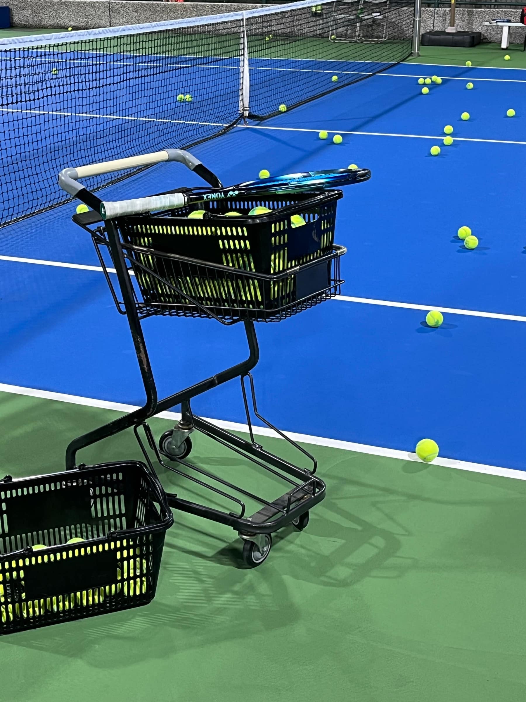 A ball cart and scattered tennis balls on a blue court with a green apron